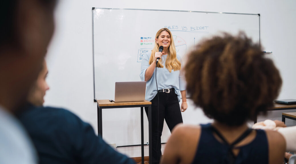 Frau steht mit Mikrofon vor Whiteboard und Laptop, Publikum im Vordergrund unscharf zu sehen.